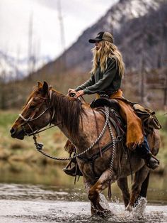 a woman riding on the back of a brown horse across a river with mountains in the background