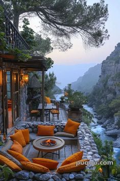 an outdoor seating area with orange pillows and chairs next to a river at dusk or dawn