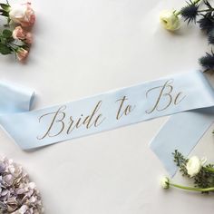 the bride to be sash is laying on top of flowers and blue ribbon with gold lettering