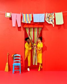 two people standing in front of a red wall with colorful clothes hanging on the line