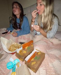 two women sitting on a bed eating french fries and macaroni and cheese sandwiches