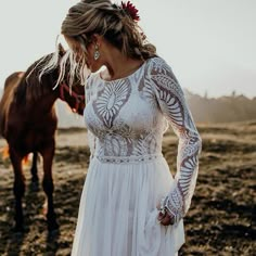 a woman in a white dress standing next to a brown horse on a dirt field