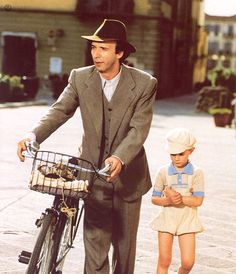 a man in a suit and hat riding a bike with a little boy standing next to him