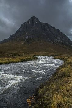 a river running through a lush green field next to a tall mountain under a cloudy sky