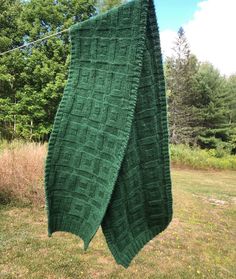 a green knitted blanket hanging from a clothes line in the grass with trees in the background