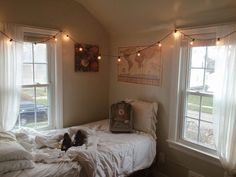 a bed with white sheets and lights hanging from the ceiling in a bedroom next to two windows