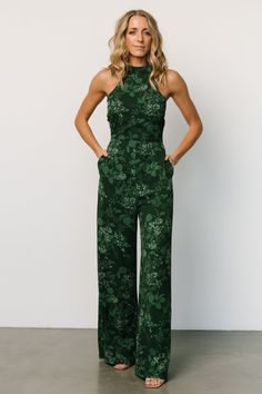 a woman standing in front of a white wall wearing a green floral jumpsuit with her hands on her hips