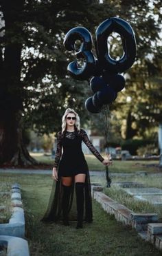 a woman standing in the grass holding some black balloons and wearing thigh high boots with her legs crossed