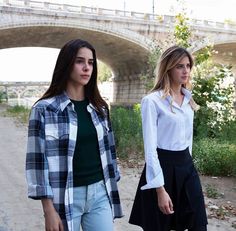 two young women walking down a dirt road under a stone bridge in the background, one is wearing a white shirt and black pants