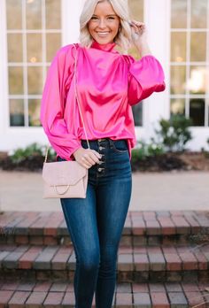 a blonde woman wearing jeans and a pink top with her hand on her hip, smiling at the camera