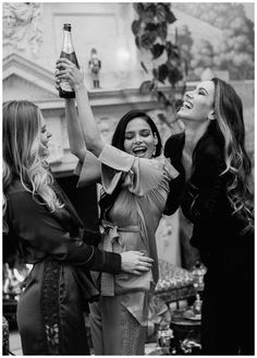 three women are holding up a beer bottle in the air and laughing while standing next to each other