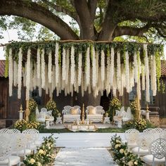 an outdoor ceremony setup with white chairs and flowers hanging from the tree overflowing