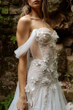 a woman in a white wedding dress standing next to a stone wall with flowers on it