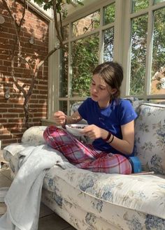 a woman sitting on a couch eating food from a bowl in her hand while looking out the window