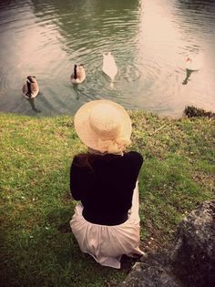 a woman sitting on the grass looking at swans swimming in a pond with their backs turned to the camera