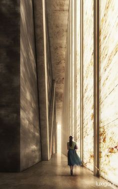 a woman in a long dress walking down an empty hallway between two tall buildings, with sunlight streaming through the windows