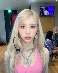 a woman with blonde hair wearing a pink top and necklaces on her neck is looking at the camera