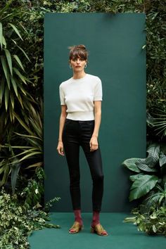 a woman in white shirt and black pants standing next to green wall with plants behind her