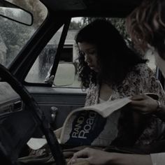 two women sitting in the driver's seat of a car, one reading a book