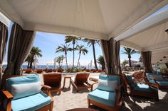 chairs and tables are set up under an awning on the beach with palm trees in the background