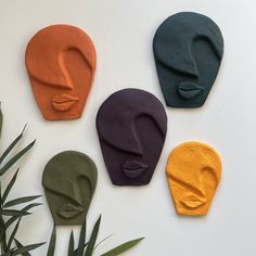 four different colored clay masks sitting on top of a white table next to a plant