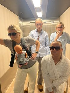 four people standing in an elevator with one holding a baby doll and the other looking at the camera