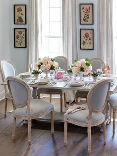 a dining room table is set with flowers and plates in front of the window, along with white chairs