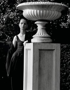 a woman standing next to a tall white vase on top of a cement pillar in front of trees