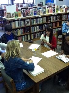 several children are sitting at a table in the library and writing on their notebooks