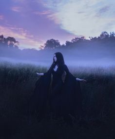 a woman with long black hair is standing in the grass at dusk wearing a cloak