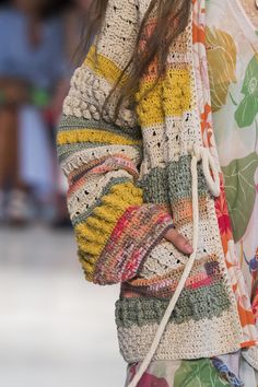 a woman walking down a runway carrying a handbag in her right hand and wearing a multicolored sweater