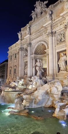 a fountain in front of a building with statues and lights on it at night time