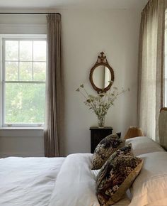 a bed with white sheets and pillows in a bedroom next to two large windows that have drapes on them