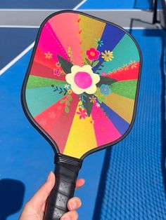 a person holding up a colorful tennis racket on a blue and black court with flowers