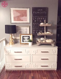 a white dresser topped with lots of drawers next to a lamp and pictures on the wall