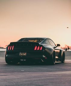 the rear end of a black sports car parked on top of a parking lot at sunset