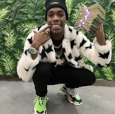 a man sitting on the ground with money in his hand and wearing a hat, fur coat, and green sneakers