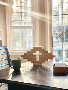 a wooden cross sitting on top of a table next to a cup and book in front of a window