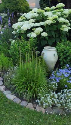 a garden filled with lots of flowers and plants next to a white vase on top of a stone path