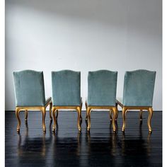 four blue velvet chairs with gold trim and wood legs on a black wooden floor in front of a white wall