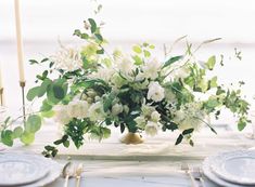 white flowers and greenery are arranged in a vase on a table set for dinner