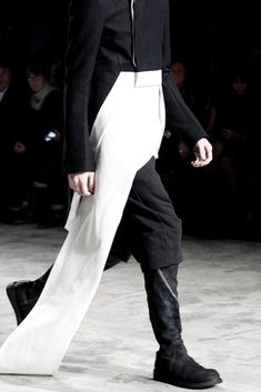 a man is walking down the runway wearing black and white pants with high boots on his feet