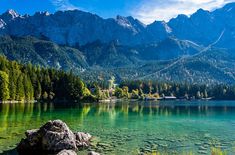 a lake surrounded by mountains and trees with clear water in the foreground, on a sunny day