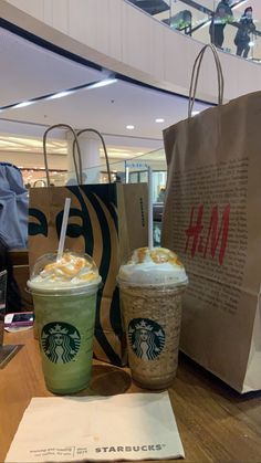 two starbucks drinks are sitting next to each other on the table in front of some paper bags