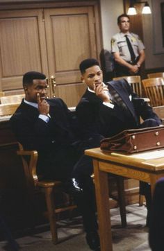 two men in suits sitting at a table talking on their cell phones while another man stands behind them