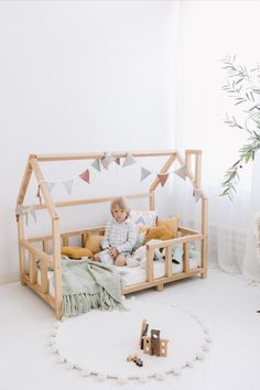 a small child sitting in a wooden bed with pillows and toys on the floor next to it