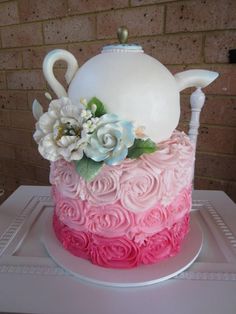 a pink and white cake with flowers on top is sitting on a table in front of a brick wall