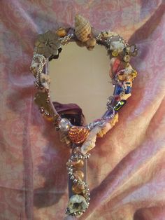 a heart shaped mirror with seashells and shells on it's sides, sitting on a pink fabric