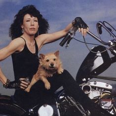 a woman sitting on a motorcycle holding a small dog and wearing leather gloves with her hand on the handlebars