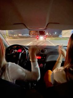 two women driving down the road at night with their hands in the air and one woman holding her head up